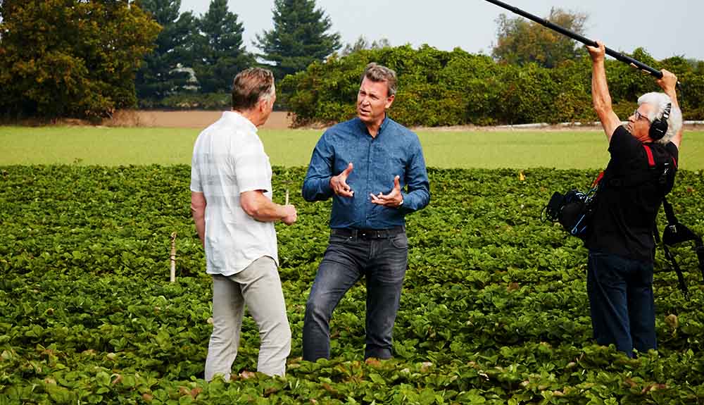 Celebrity chef Ricardo Larrivee chats with a man in a field while a boom operator stands capturing audio nearby