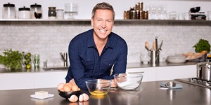 Celebrity chef Ricardo Larrivee smiles and leans on kitchen counter, flour and eggs in a bowl in front of him