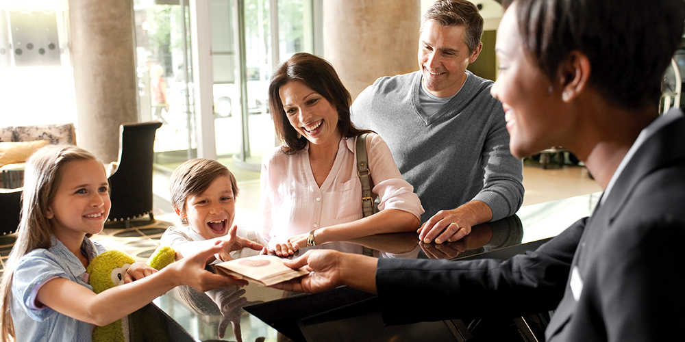 Mom, dad and two young children, all with happy, excited faces, reach for a brochure packet from a hotel concierge