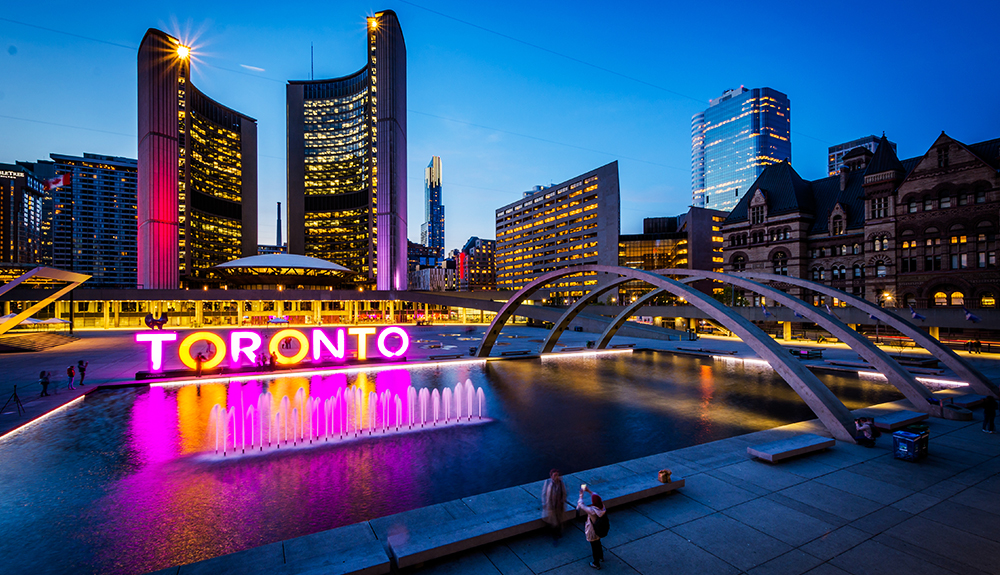 Toronto city hall area seen at dusk, the bright pink and orange lights of a Toronto sign reflecting in a small pond and the glittering lights of high rise buildings seen in the background