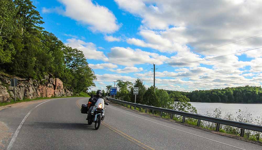 A black motorcycle drives along a winding road alongside a lake under a beautiful blue sky