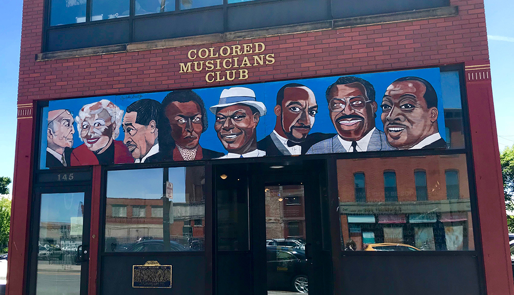 Brick building identified as the Colored Musicians Club, seen with various famous Black musicians painted on the sign