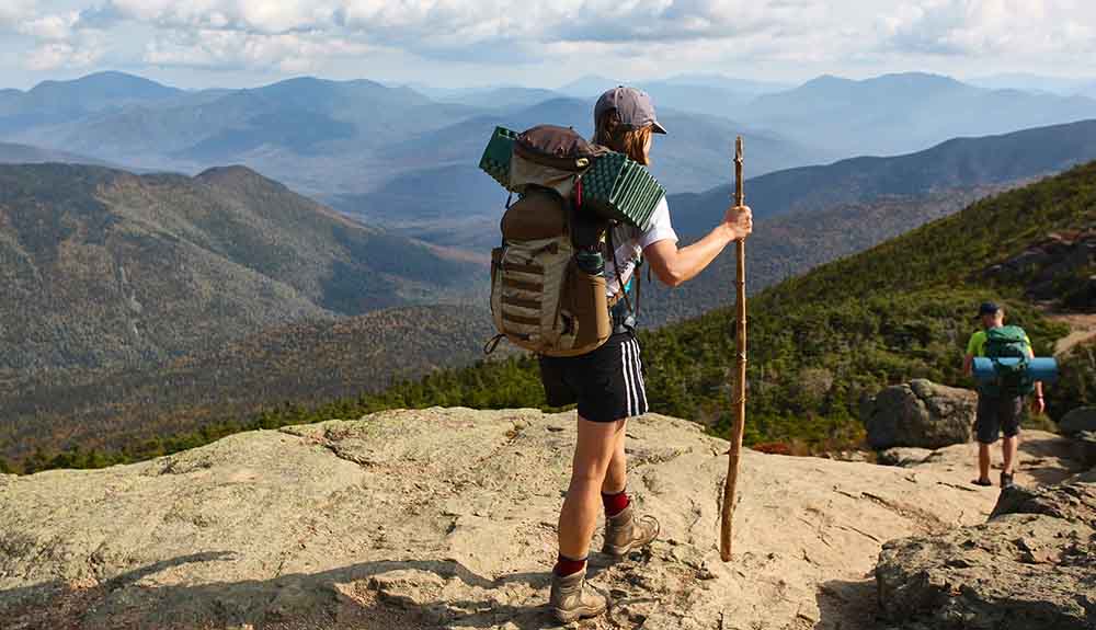 Two people seen hiking a rocky, mountainous path with hiking sticks and large packs on their backs in the White Mountain National Forest trail