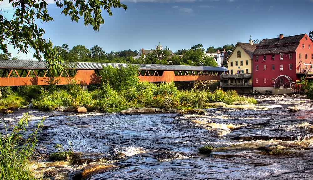 Lively river splashes in front of an orange covered bridge leading to small, colourful homes