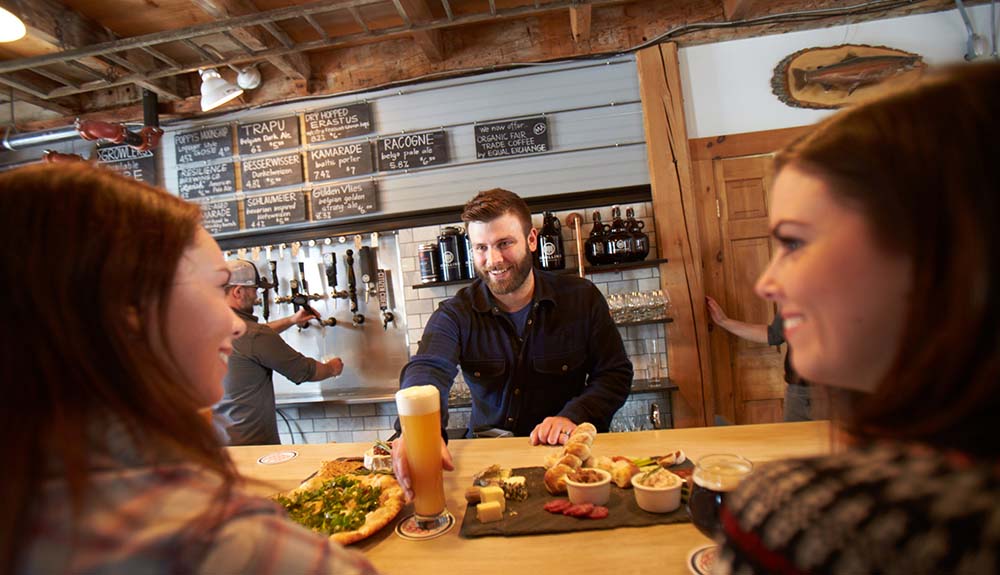 Two young women being served a pizza, charcuterie plate and large mug of beer at a New Hampshire brewery