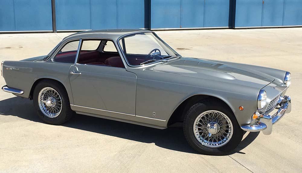 Slate grey 1960 Triumph Italia 2000 GT Vignale Bodied car parked on light driveway in front of blue building