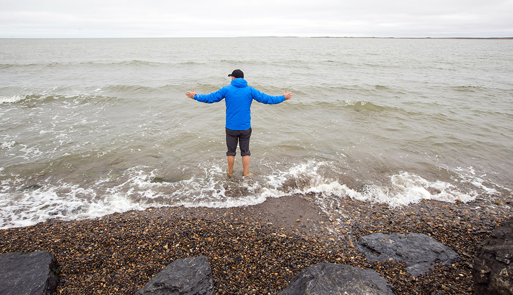 A young man in blue coat and black cap is seen wading in the gentle waves of the ocean on a rocky shoreline