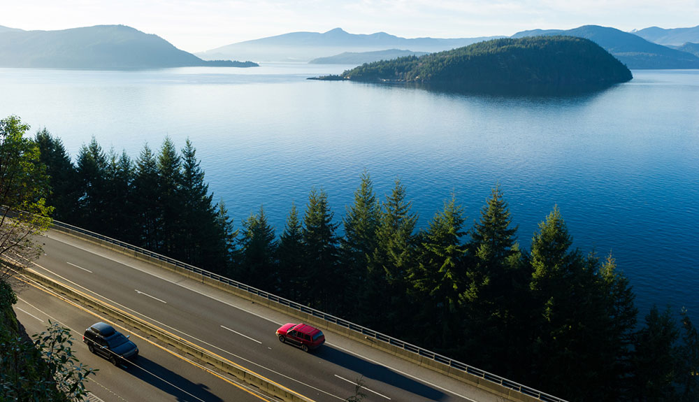 Overhead shot of Route 99 highway in British Columbia, two cars seen driving in opposite directions beside the beautiful blue ocean