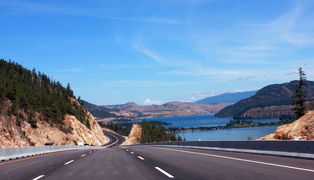The expansive winding road of Route 97 in British Columbia is seen with no a car in site, mountains lined with trees on one side and bright blue water to the other