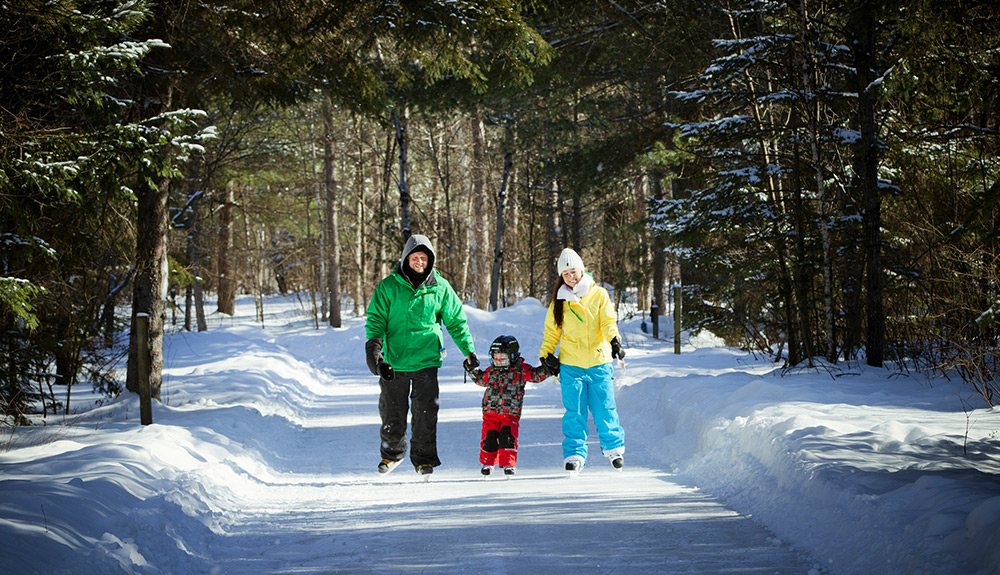 Mom, dad and child skate along an iced pathway wearing bright warm winter wear in tree-lined Ontario cottage country