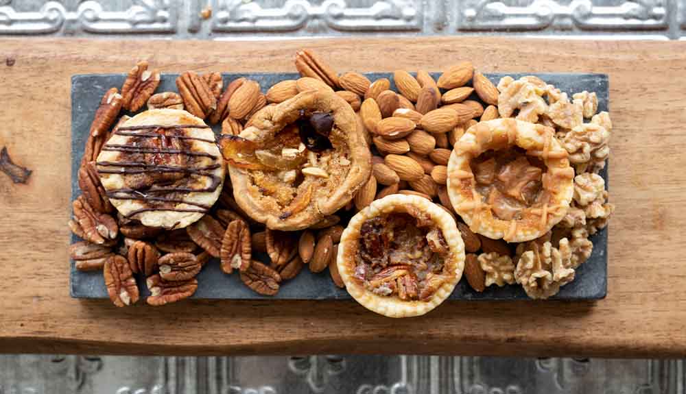 An overhead photo shows four butter tarts on a wood board with almonds, pecans and walnuts
