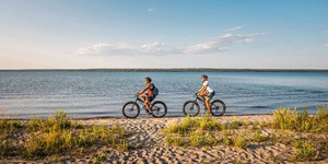 Two adventurous cyclists navigate a sandy trail along a serene beach or lake shoreline, with tufts of green grass punctuating the sand. A vivid blue sky overhead, lightly speckled with wisps of clouds, adds to the tranquility of a bright, sunny day.