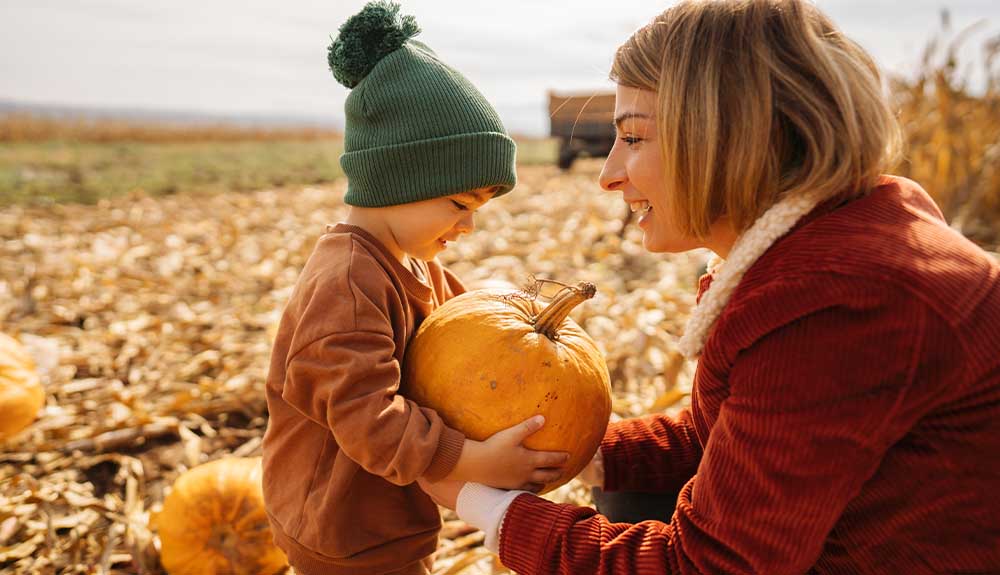 A woman with shoulder-length blonde hair wearing a copper-coloured corduroy coat with a shearling collar is facing a young child wearing a brown sweatshirt and an olive green toque. He is holding a small pumpkin with both of his hands.