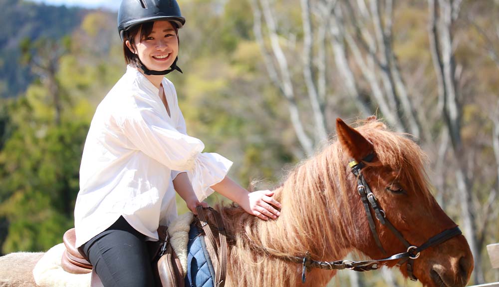 A woman wearing a white billowing blouse and black pants is sitting on top of a chestnut-coloured horse. She is wearing a black helmet and patting the horse's mane.