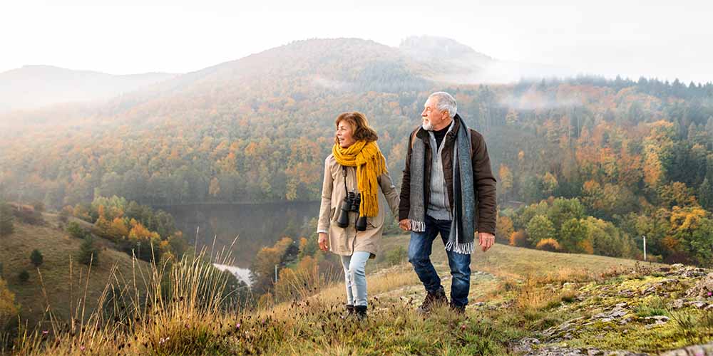 A man with silver hair wearing a brown coat with a scarf around his shoulders, jeans and a grey sweater is holding hands with a woman. She is wearing a long beige coat with a gold-coloured scarf around her shoulders. There is a pair of black binoculars dangling from her neck. They are walking on a green hill with trees in shades of green and orange behind them.