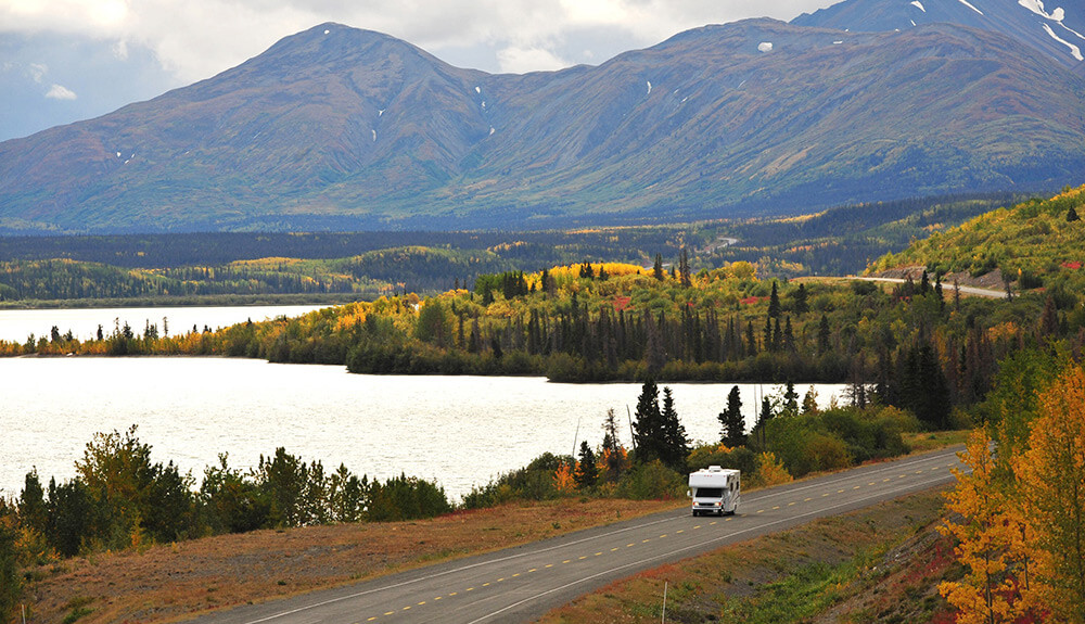 Fall leaves colour the scene as a white RV drives along the picturesque Dempster Highway beside a body of water, mountains seen in the background