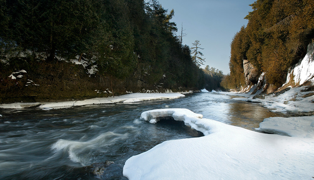Closeup shot of an icy winter river covered with snow in some patches, the riverside lined with tall trees