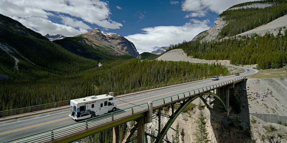 A white RV drives along an elevated bridge down a winding highway path, snow-topped mountains seen on every side