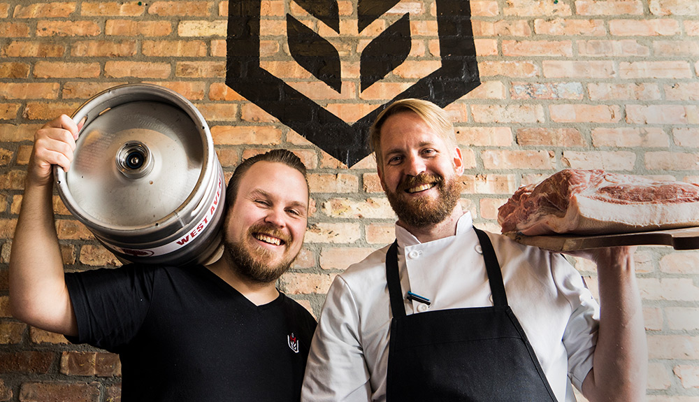 Two men stand in front of brick wall, one holding a cask of beer, one holding a wooden cutting board with large cuts of raw steaks