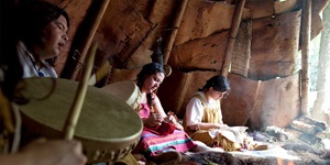 Four Indigenous people sit in a teepee. From the left: a drum and drumstick being played, a man with dark brown hair and white shawl singing and playing a small drum, a woman with brown hair in two braids, wearing pink and handling a red garment, a young woman with brown hair in a ponytail, wearing a white blouse and mustard overalls with fringe and moccasins, handling a white garment. There is sunlight streaming in from outside from an opening on the right.