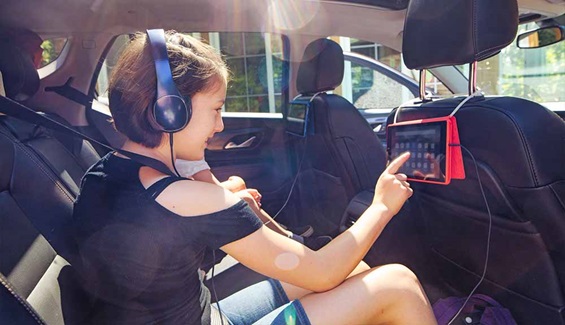 A child is shown in the backseat of a car wearing headphones and playing on a tablet
