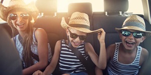 Three kids wearing sunglasses and straw hats are shown sitting in the backseat of a car