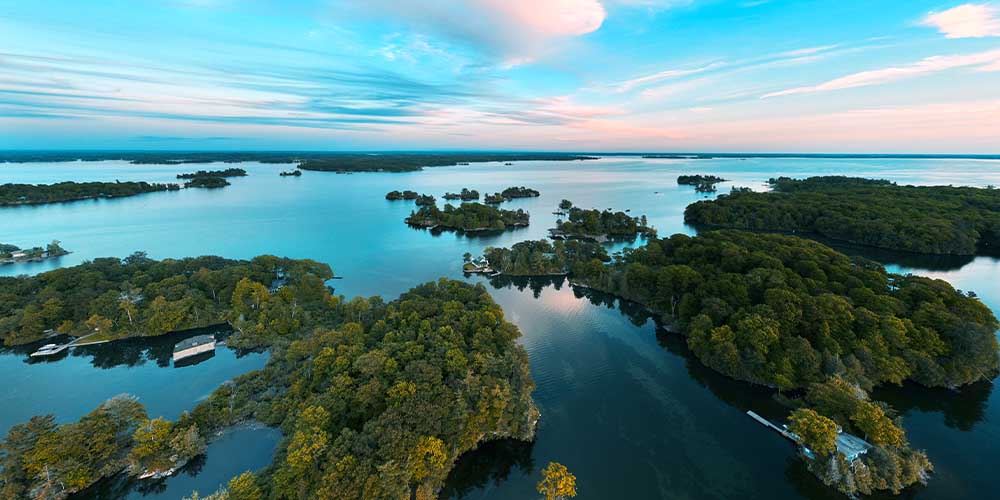 Aerial view of the 1000 Islands in Kingston, Ont. You see lush greenery and trees on the islands, which are scattered across a blue body of water. The sky is light blue with white clouds, which is reflected in the water.