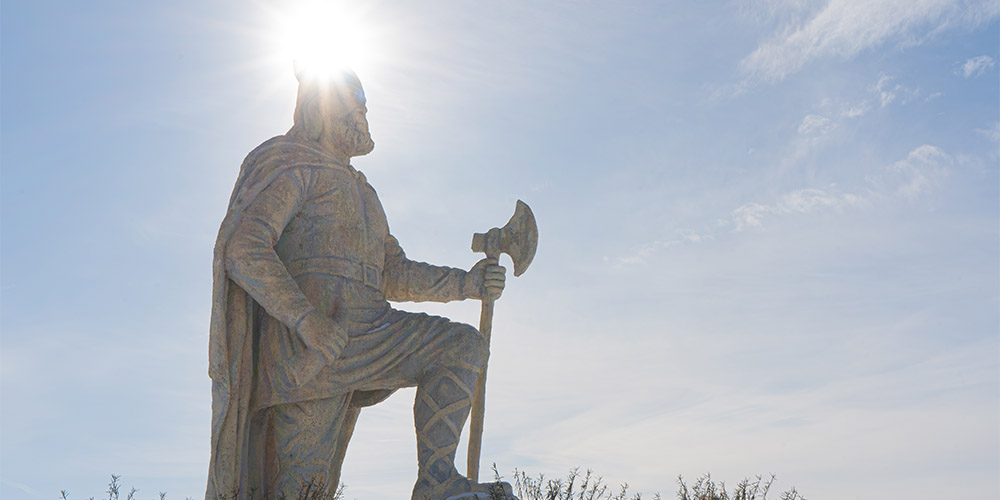 A large, tall statue of a gladiator figure wearing a helmet, cloak, shirt, belt, pants and sandals with a strap wrapping around his shin to his knee. He is holding a long axe in front of him. You see the sun behind his head and the sky is blue.