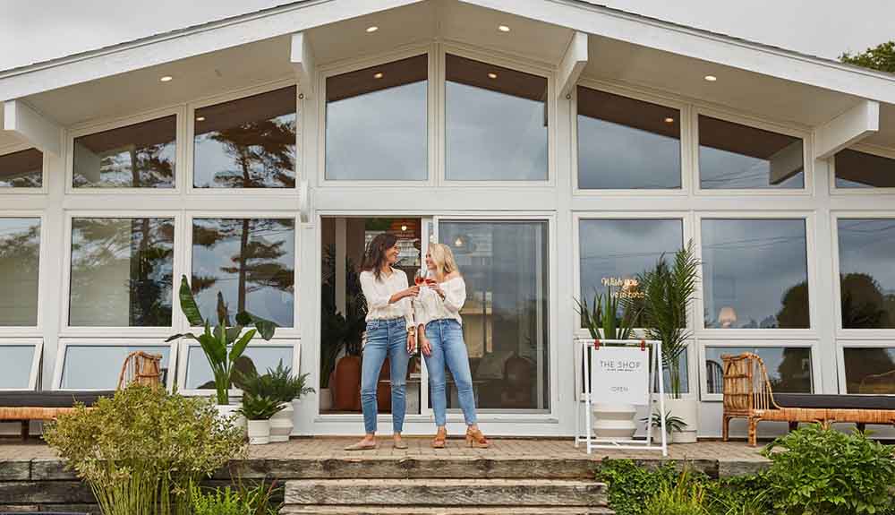 Two women wearing white shirts and jeans toasting with glasses of red wine standing on a stone deck. Behind them is a building covered with windows.
