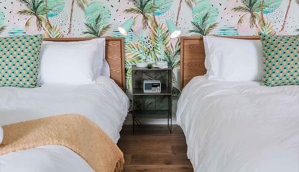 Two beds with teak headboards are side by side. Each bed is made up with white bedding and a decorative throw with a pineapple print on it. There is a tropical-themed wallpaper on the wall behind the beds.