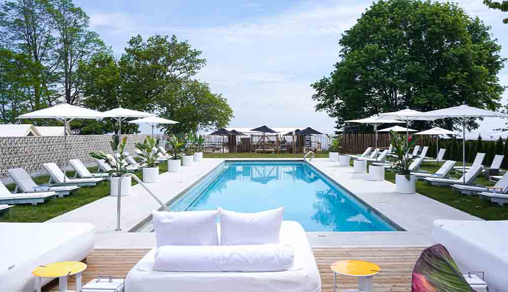 A pool is in the centre surrounded by white deck chairs and umbrellas on the perimeter. There is a large tree to the right of the pool.