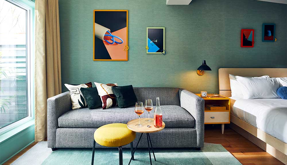 A grey upholstered couch is positioned between a window and a bed. There is a small side table in front of the couch with an open bottle of wine and two glasses with wine in it. There are colourful picture frames on the wall with abstract pieces of art in them.