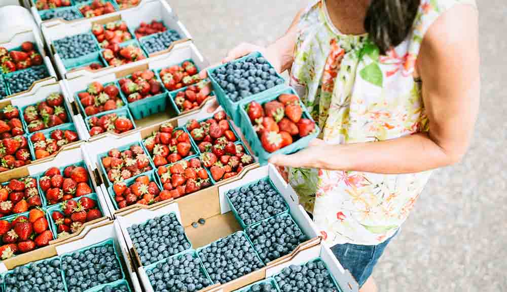 A woman in a light floral print sleeveless top is standing in front of a fruit stand displayed with pints of strawberries and blueberries. In her hand she holds a pint of blueberries and a pint of strawberries next to each other.