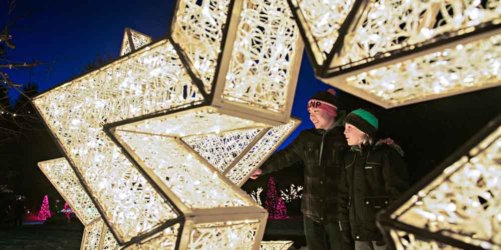 Two people stand in front of a large festive white and gold weaved star. You can see them through the center of the star, one wearing a red and black beanie with a pop-pom, a plaid green coat and black pants. They are smiling and reaching out toward the star. The second stands close, wearing a green and black hat with a pom-pom, a black coat and gray pants.