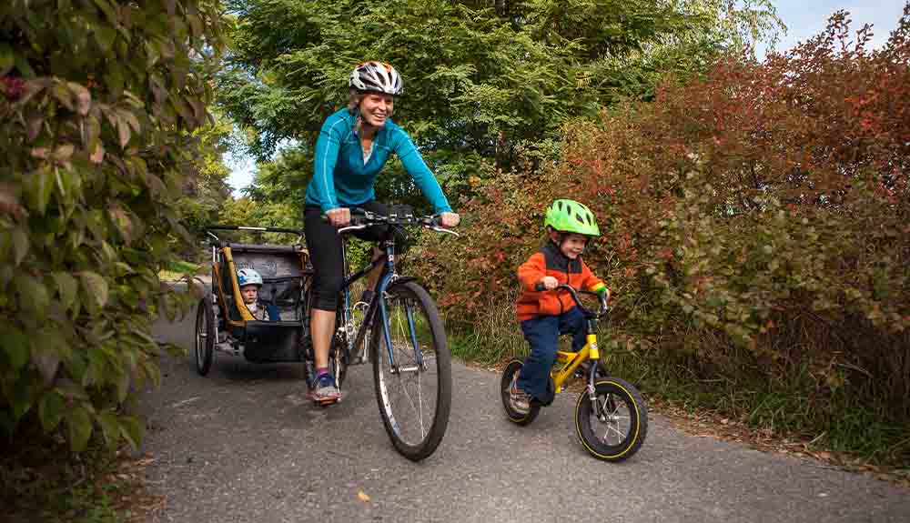 A parent is shown cycling with a bike trailer behind her and a young child riding a bike next to her on a path through trees