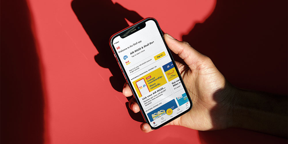 A hand holds a smartphone displaying the Shell app, which features various offers.