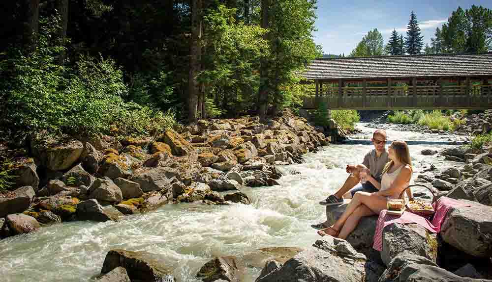A couple enjoys a picnic next to a river on the property of the Fairmont Chateau Whistler