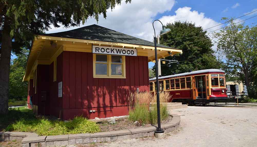 A small burgundy building with yellow paint under the roof and a black and white sign that says Rockwood is in one corner. Behind it is an old-fashioned TTC streetcar in the same shade of burgundy accents with yellow trim around the windows and doors.