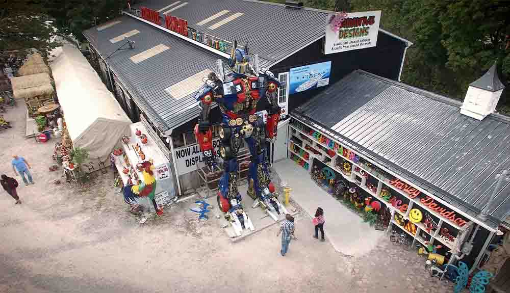 An overhead view of a store. In the middle is a blue, red and black robot figure that is taller than the building. There are three people standing on the ground looking up at it. To the right of them are retro looking signs. The right of them are other items for sale, like a colourful rooster.