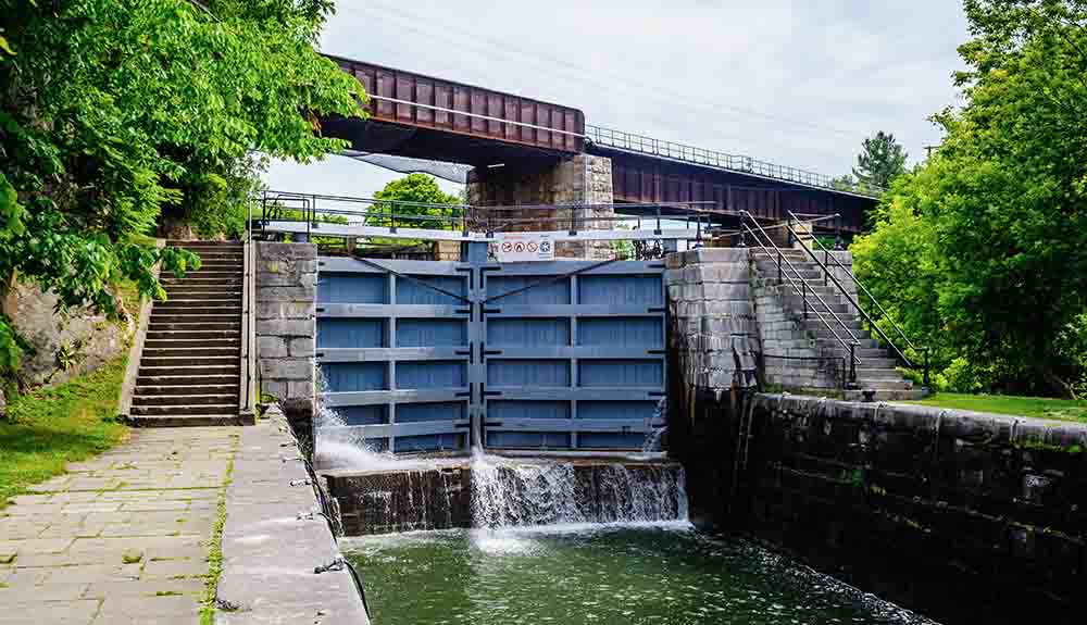 A bridge overhead  and below it, a small dam with blue gates over it, and two flights of concrete steps next to it with a walkway.