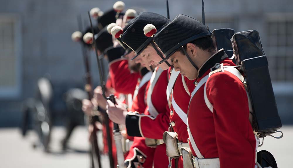 A row of soldiers standing in a line. They are dressed in red jackets with bands of white criss crossing their chest and a white belt. They are wearing black hats with a ball that is half white on top and half red on the bottom.