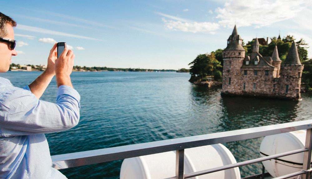 A man holding a cellphone up to take a photo of a stone building with turrets in the distance. He is on a boat, surrounded by water.
