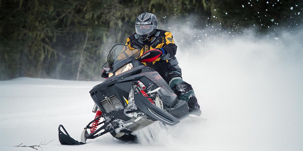 A person wearing an orange and black winter jacket, black helmet and black snow pants rides a black snowmobile in a snow-covered field lined with evergreen trees. Snow sprays from behind it.