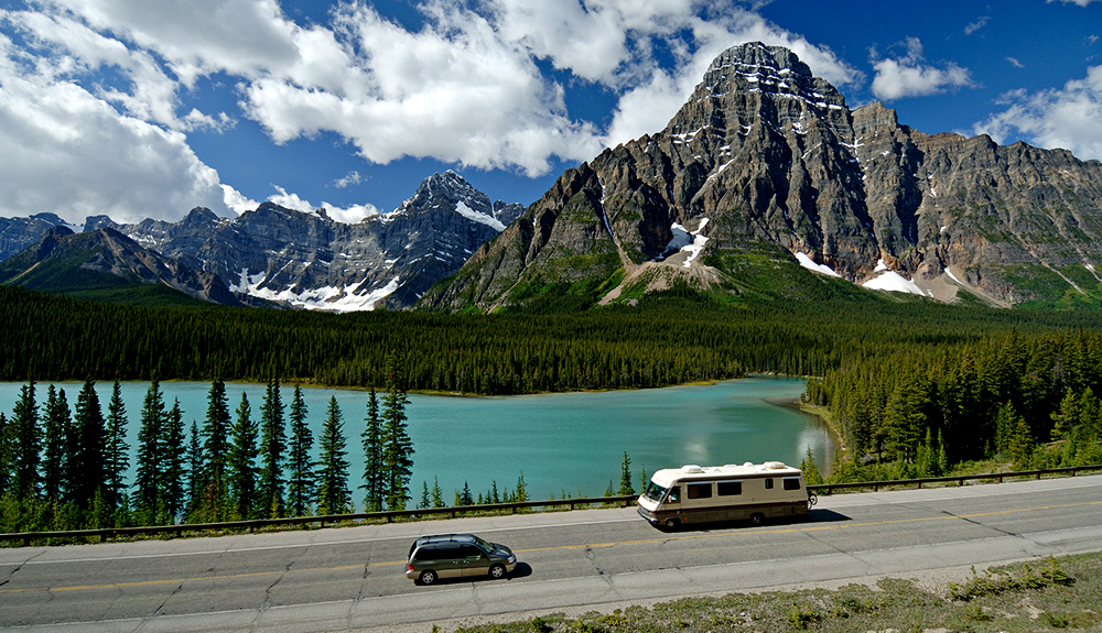 An RV and van are seen passing in opposite directions on the roads in front of a beautiful lake before the Canadian Rockies