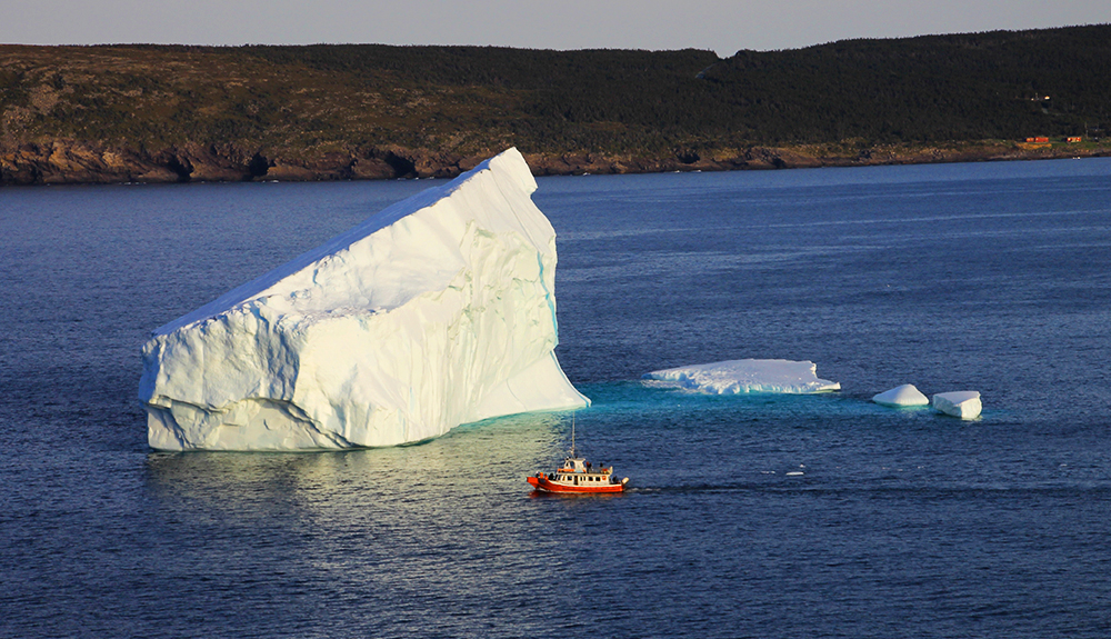 A large iceberg is seen in the ocean, a small red ship nearby