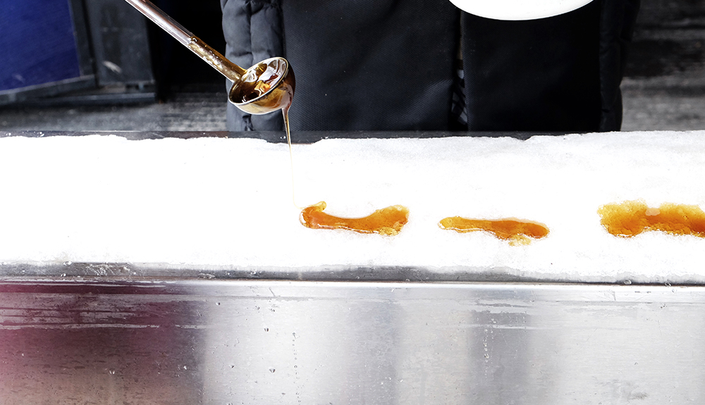 Hot maple syrup is poured from a ladle onto fresh white snow