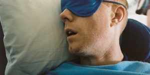 A man cozied up in a blue fleece blanket sleeps against a grey pillow, a soft blue eye mask cover his eyes