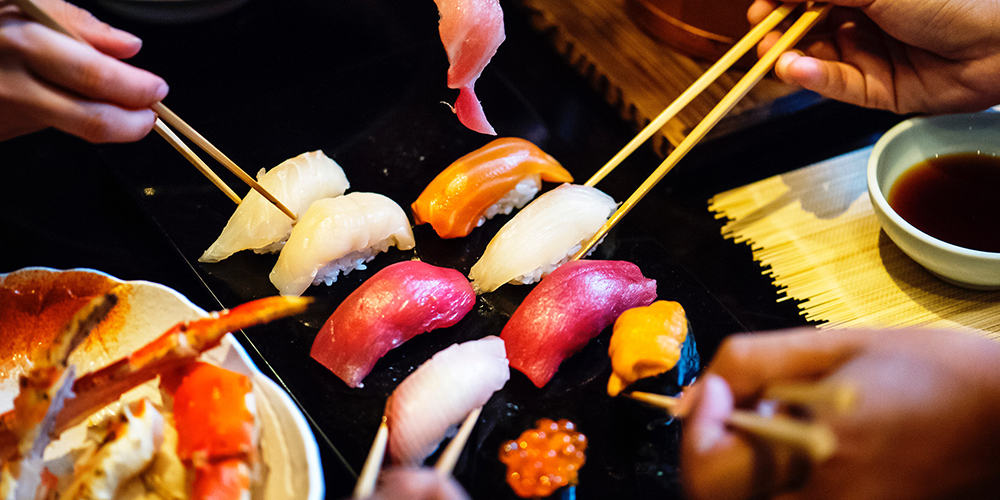Fresh seafood nigiri laid out, several hands with chopsticks reaching for a piece