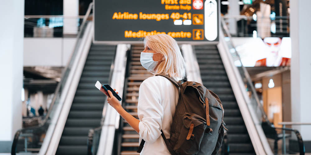 A profile view of a blonde haired woman standing in front of two escalators, wearing a light blue mask and a white sweater with the sleeves pushed up to the elbow and a grey backyard with brown straps. She is holding a cell phone and passport with a boarding ticket inserted inside in her left hand. 