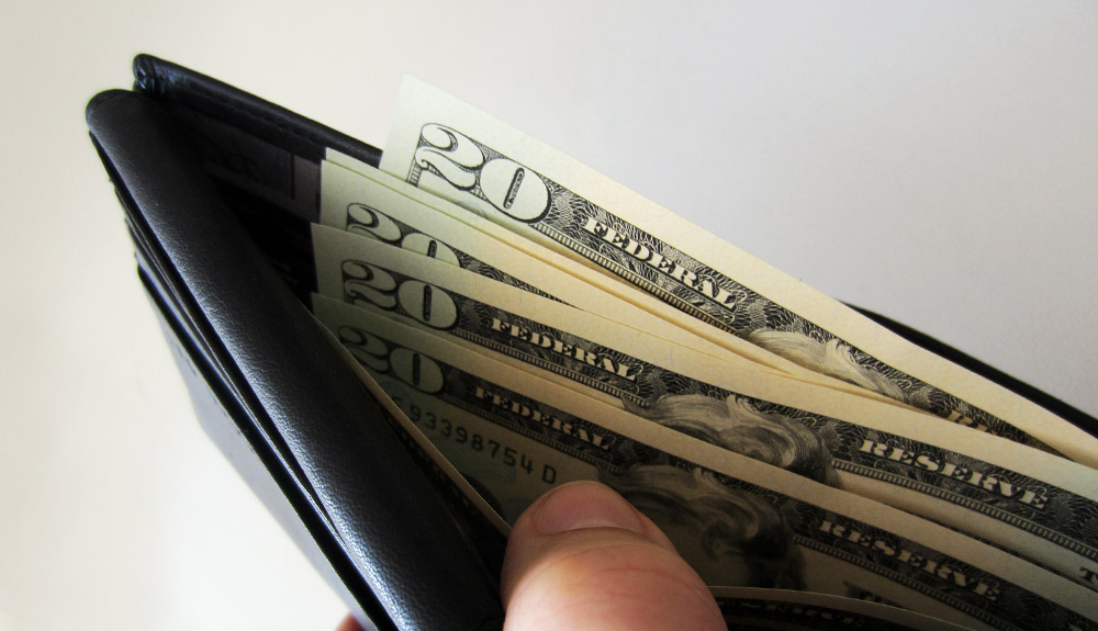 A hand opens the pocket of a black wallet, a stack of American twenty dollar bills inside